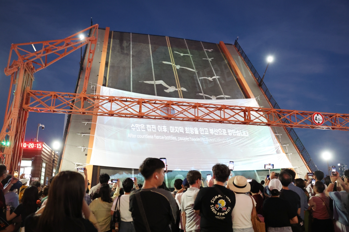 ▲7月25日，釜山市民参加在釜山中区影岛大桥举行的“2026韩国战争临时首都釜山国家遗产夜游”活动，欣赏夜间表演，并观看讲述釜山作为韩国战争期间临时首都、历时1023天的历史主题影像。影岛大桥始建于1932年，是韩国首座连接陆地与岛屿的跨海桥梁，连接釜山中区与影岛区。韩国战争期间，这里见证了无数离散与重逢，承载着人们的悲欢离合，成为战争时期临时首都釜山最具代表性的历史地标之一。韩宣网记者朴大珍 摄